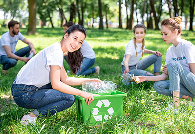 A group of people wearing casual clothing are outdoors in a park, engaged in a recycling activity. One person in the foreground places plastic bottles into a green bin with a recycling symbol