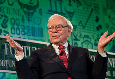 An older man in a suit and tie gestures with both hands while speaking at an event, reminiscent of Warren Buffett's style