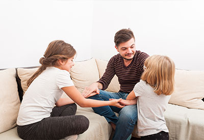 A man sits on a couch holding hands with two young girls, embodying the warmth of blended families. One girl, with long hair tied back, sits beside him, while the other, with shorter hair, faces him from the opposite side