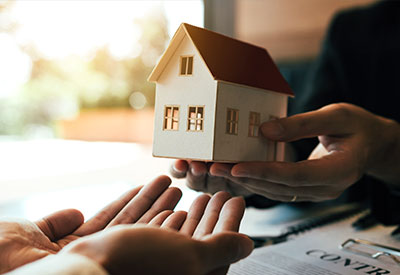 Two people exchange a small house model over a contract and pen, symbolizing estate planning or a real estate transaction