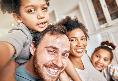 A cheerful blended family of four poses indoors, with two children on the adults' shoulders.