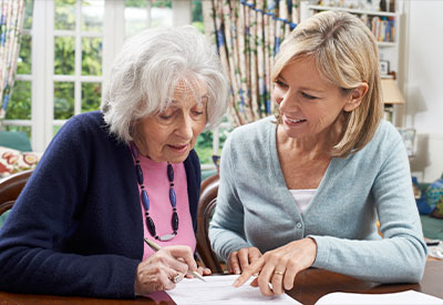 An older woman with gray hair and a pink top is writing about estate planning on a piece of paper, while a younger blonde woman smiles and points at the paper. They are seated in a cozy room with patterned curtains and a window.