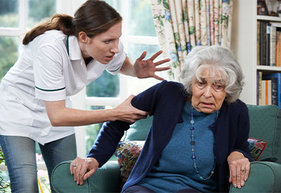 A caregiver in a white uniform appears to be speaking sternly to an elderly woman sitting in a green armchair.