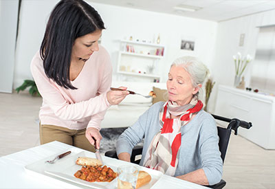 A woman, embodying guardianship, assists an elderly lady in a wheelchair by feeding her at a table.