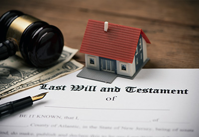 Image of a small model house placed on U.S. dollar bills near a gavel and a 'Last Will and Testament' document. A fountain pen rests on the document, highlighting legal and financial matters related to inheritance laws and estate planning for blended families.