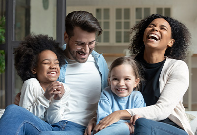 A joyful family of four sits together on a couch, laughing. Two adults and two children share a moment of happiness, dressed casually, with bright expressions