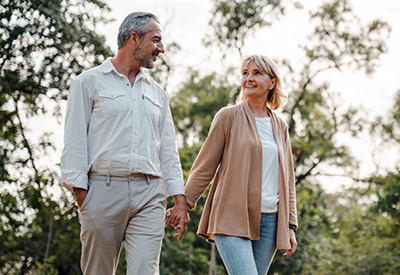 A middle-aged couple, embodying the essence of modern families, walks hand in hand through a park. The man wears a light-colored shirt and pants, while the woman dons a white top and beige cardigan.