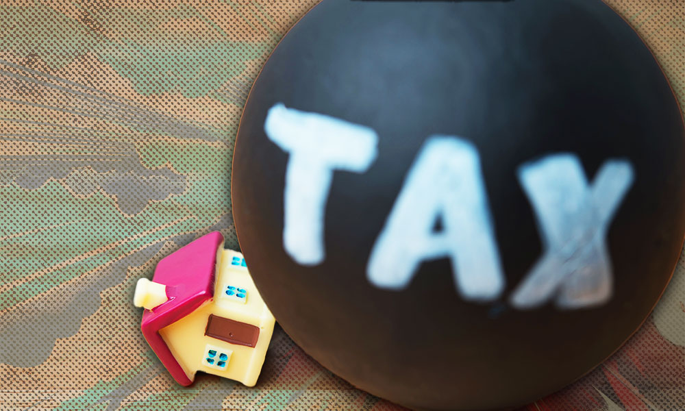 A toy house crushed beneath a heavy black ball labeled 'TAX'