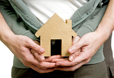 Two pairs of hands gently hold a small wooden house model, symbolizing themes of home ownership and protection