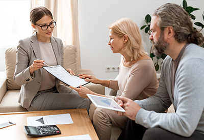 A woman in a suit holds a clipboard and explains estate planning to a seated middle-aged couple in a living room