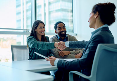 A woman and a man sit at a table in an office, both smiling as the woman shakes hands with another in a business suit, possibly discussing estate planning