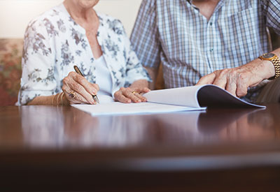 Two elderly individuals sit at a table, reviewing paperwork together. One writes as the other gestures, discussing an advance directive
