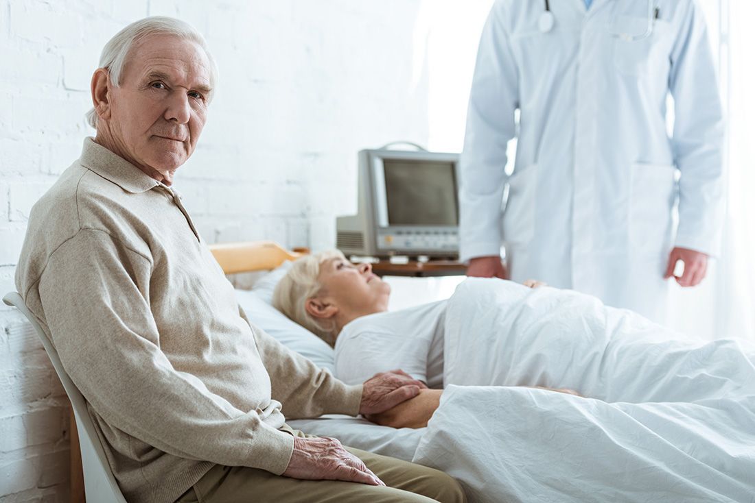 a male senior sitting beside his significant other laying in a hospital bed
