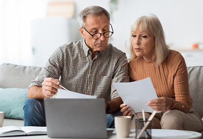 An older couple sits on a couch, focused on estate planning documents in their hands. The man wears glasses and a checkered shirt, while the woman wears a light brown sweater. A laptop and coffee mugs are on the table in front of them.
