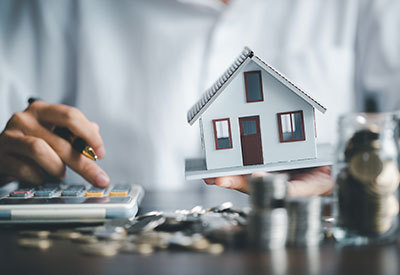 person at desk calculating finances while holding a miniature house in one hand