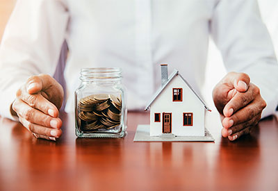 A person in a white shirt places their hands around a small glass jar filled with coins and a miniature house model on a wooden table