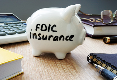 A white piggy bank with 'FDIC insurance' written on it sits on a wooden desk, surrounded by tools for estate planning—a calculator, notebooks, a pen, and a pair of eyeglasses