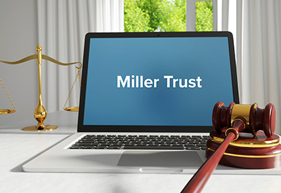  laptop displaying 'Miller Trust' alongside a gavel and scales of justice represents the legal foundation of Qualified Income Trusts for Medicaid eligibility.