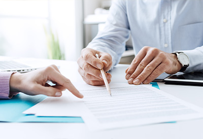 Two individuals sitting at a table reviewing documents for estate planning. One person is holding a pen pointed at the text, while the other points with a finger.