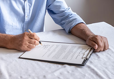 A person in a light blue shirt is seated at a table, signing a life insurance document on a clipboard with a pen in their right hand.