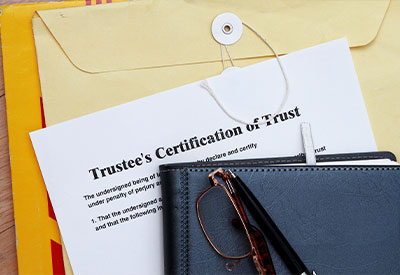 A Trustee's Certification of Trust document partially peeks out from under a closed manila envelope with a string tie. A pair of reading glasses and a pen rest on a dark leather-bound book