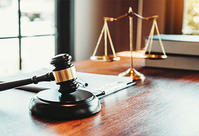 A wooden gavel rests on a table next to a pen and a stack of books—perfect tools for handling estate planning. Behind them, a brass balance scale gleams near the window.