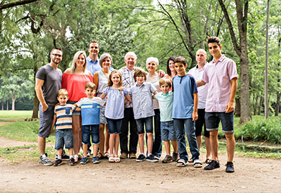 A large, multi-generational family poses together in a park. They stand in two rows, with trees and greenery in the background. 