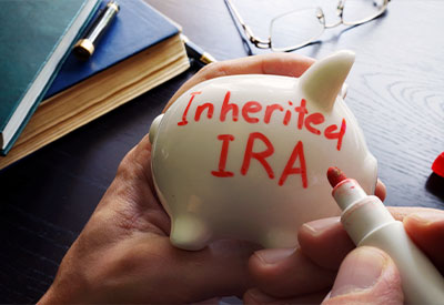 A person holds a white piggy bank labeled 'Inherited IRA' in red marker