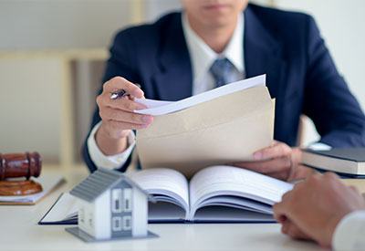 A lawyer in a suit reviews documents while sitting at a desk with an open law book, a small house model, and a gavel, indicating a focus on estate planning or property law.