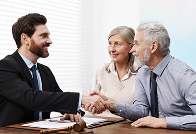 A senior couple shakes hands with a young male attorney across a desk, smiling and engaging in a positive interaction during their estate planning meeting, with documents and a gavel visible.