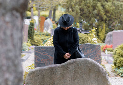 A person in black clothing and a hat stands in front of a gravestone, head bowed in quiet reflection. 
