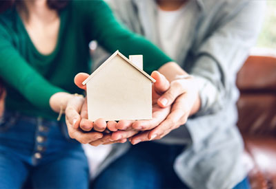 A couple, casually dressed and seated on a couch, holds a small wooden model of a house, representing their future in estate planning.