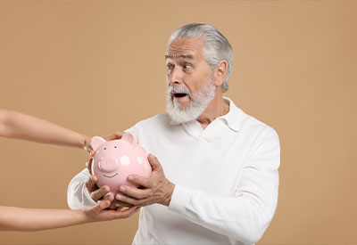 An older man with gray hair and beard, wearing a white shirt, looks surprised while holding a pink piggy bank. As another person's hands reach for it