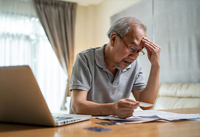 An elderly man wearing glasses sits at a table with a laptop open, holding his head in one hand and an estate planning document in the other.